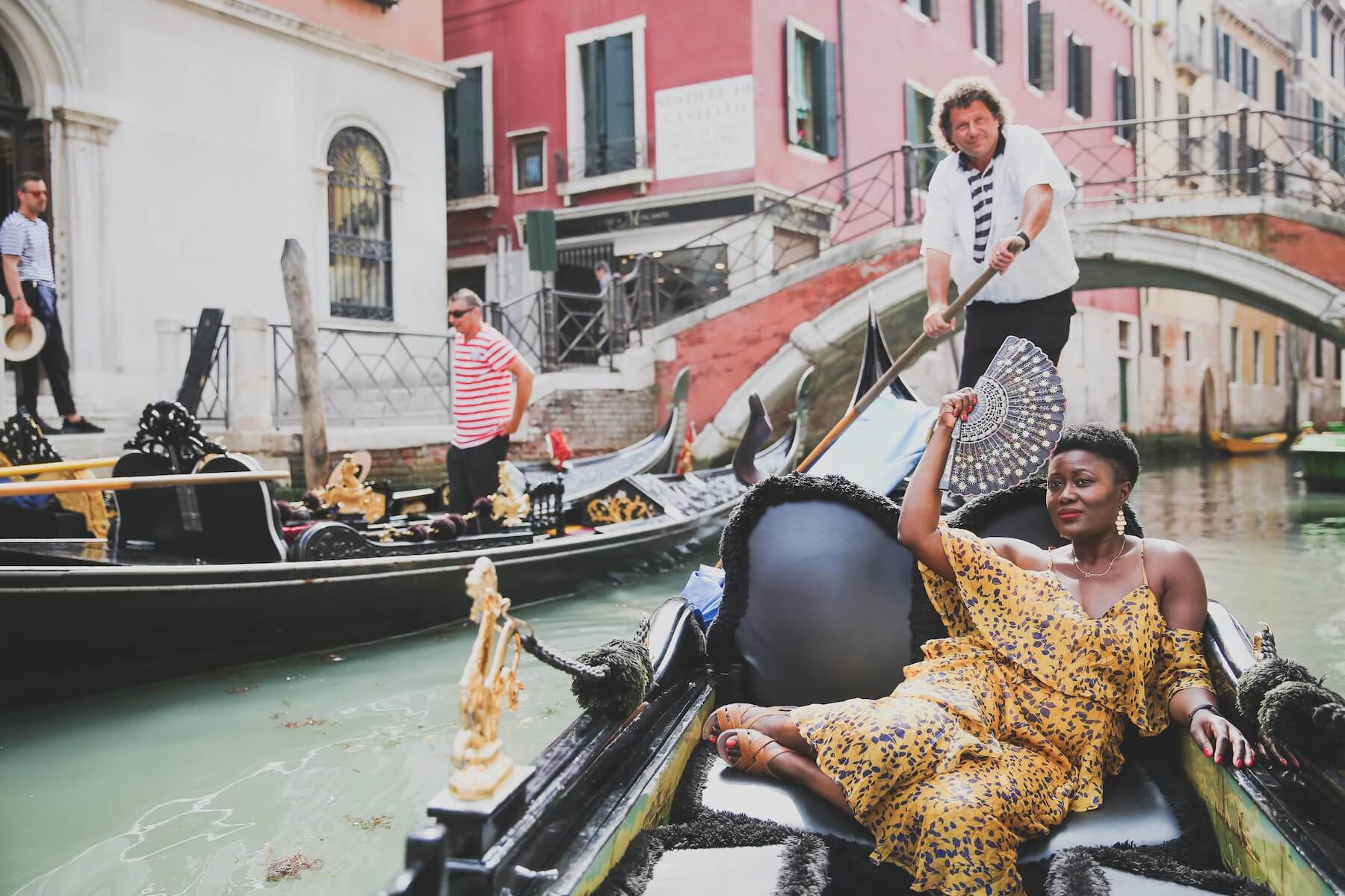 A woman riding in a gondola in Venice on a solo photoshoot with Flytographer wearing a yellow dress and holding a fan